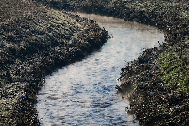 Narrow stream flowing through a frosty rural landscape in early morning light. Cold winter atmosphere with bare vegetation and gentle mist rising from the water. Natural outdoor scene with no people.