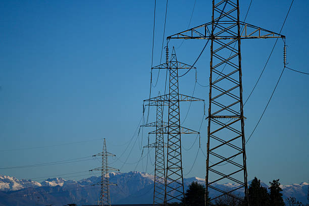 Series of high voltage electricity transmission towers and power lines stretching across the landscape, with a mountain range visible in the background under a clear sky. Scene representing energy infrastructure, electricity distribution, technology, industry, and the relationship between nature and human-built systems.