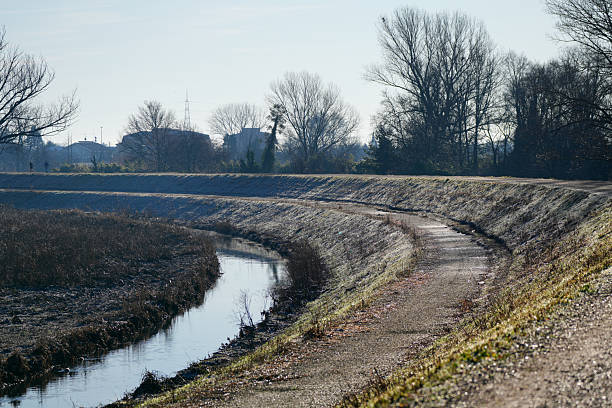Empty pedestrian path running alongside a small canal in a suburban or rural landscape, with leafless trees and embankments in winter or early spring. Outdoor scene captured in natural daylight, suitable for concepts of journey, direction, infrastructure, water management, environment, and copy space.