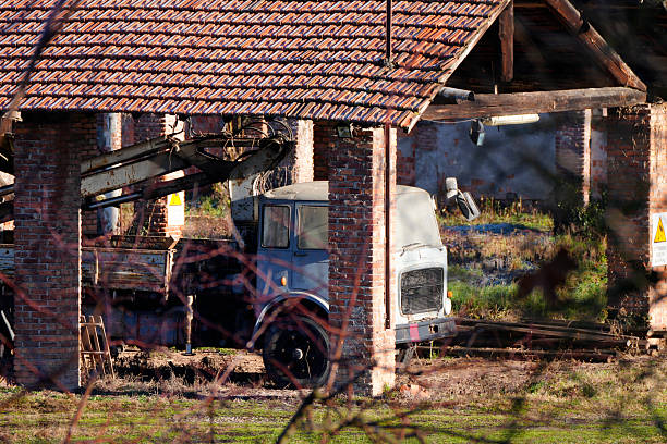 Old abandoned industrial truck parked inside a brick shed with tiled roof in a rural or industrial setting. Weathered structure and machinery surrounded by natural light and shadows, captured outdoors in daylight. Suitable for concepts of industrial heritage, decay, abandonment, history, rural industry, and transition over time.