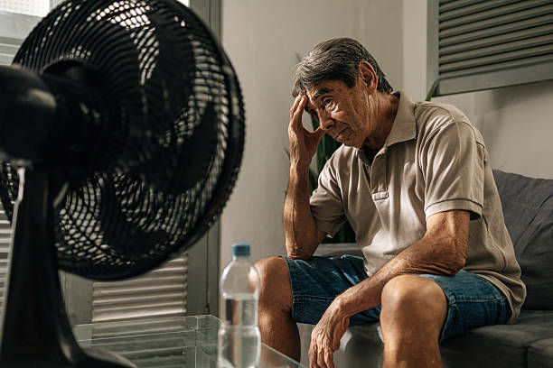 istock Senior man sits indoors feeling discomfort from heat while cooling himself with a fan, touching his head and resting, illustrating summer heat, fatigue, vulnerability, and everyday health concerns 2253829409