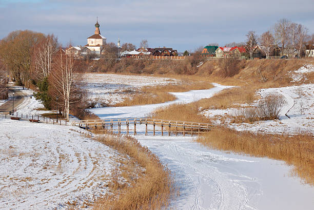 Suzdal, Russia - February 23, 2025: Serene winter view of the old city with the Kirzhach gangway bridge, the Kamenka river and wooden houses.