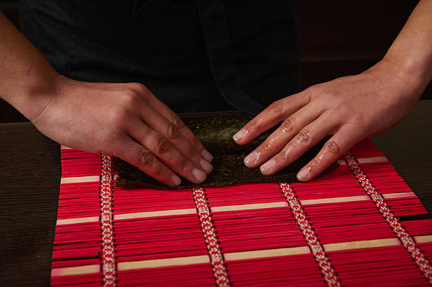 Two hands press a sheet of nori over a red bamboo mat, shaping a sushi roll. Focused culinary action captures craftsmanship and fresh ingredients in a warm kitchen setting.