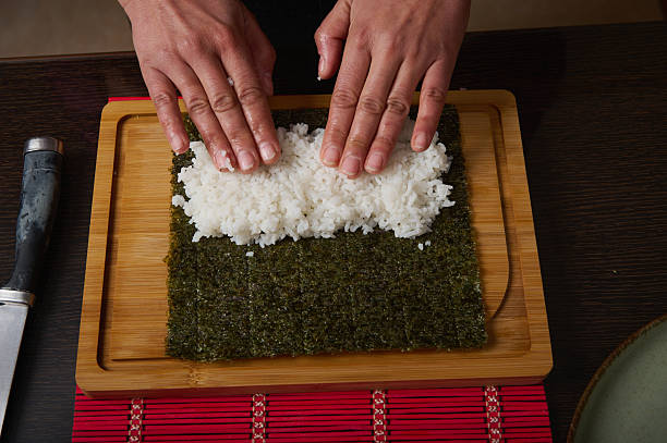 Close-up of hands spreading rice over a square sheet of seaweed, ready to roll sushi on a wooden cutting board.