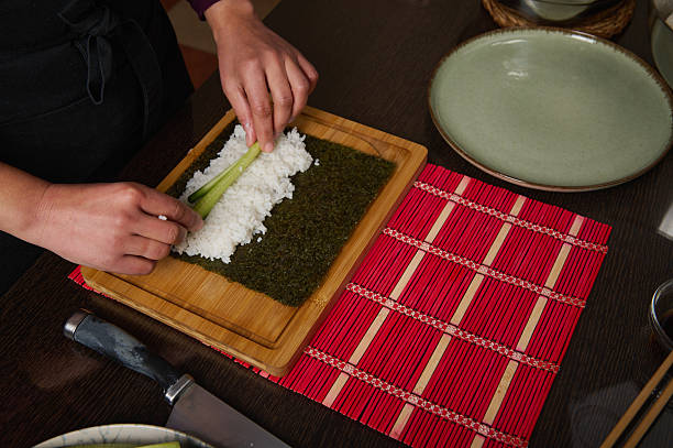 A cook rolls sushi by layering white rice on nori seaweed, using fresh leek as a filling on a wooden board beside a red placemat, capturing home-cooked Japanese cuisine.