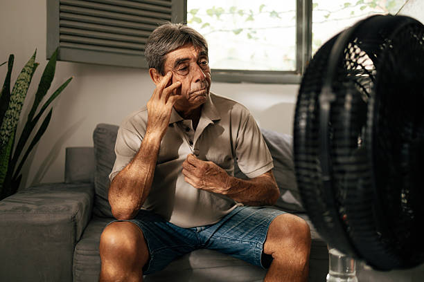 istock Senior man sits indoors feeling discomfort from heat while cooling himself with a fan, touching his head and resting, illustrating summer heat, fatigue, vulnerability, and everyday health concerns 2253834252