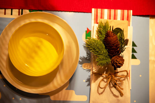 One Christmas flower in a pot stands on a festively served table with gray plates, knives, forks, glasses, mini Santa Claus hats, a red tablecloth and green tinsel on a winter evening in the kitchen, close-up top view with selective focus.