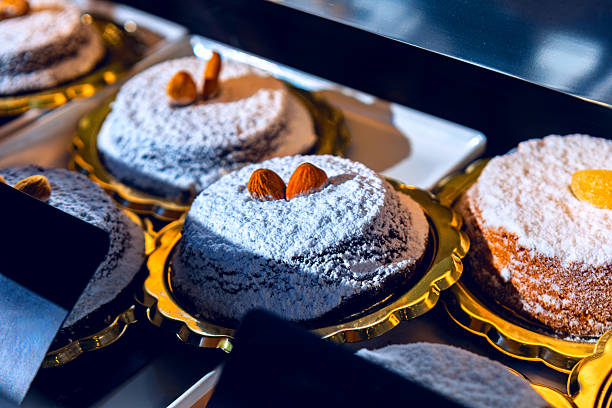 Trays cradle round Caprese cake dusted with powdered sugar, topped with almonds or candied fruit. Neat arrangement and rich texture glow under warm bakery lighting, evoking artisanal indulgence