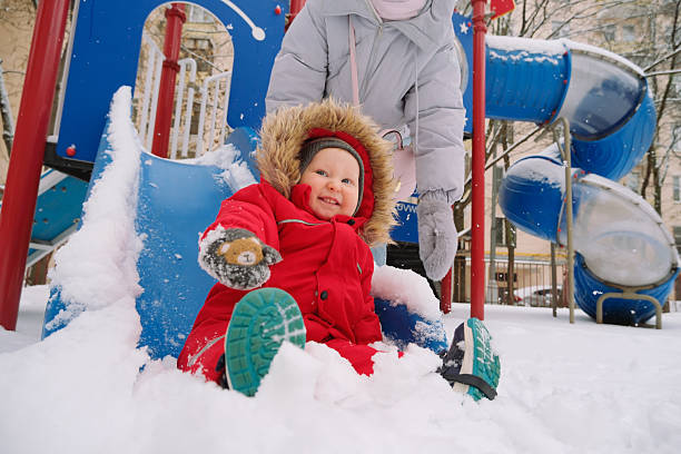istock Young child in red winter coat sliding down a snowy playground slide. Enthusiastic expression indicates excitement and enjoyment. Nearby adult ensures safety during this fun activity. 2253846265