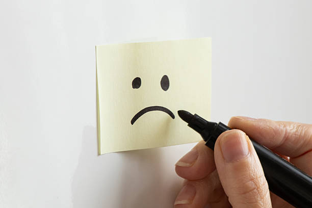 A yellow sticky note with a drawn sad face and a woman's hand holding a black marker on a white background, depicting bad mood, sadness, and negative emotions