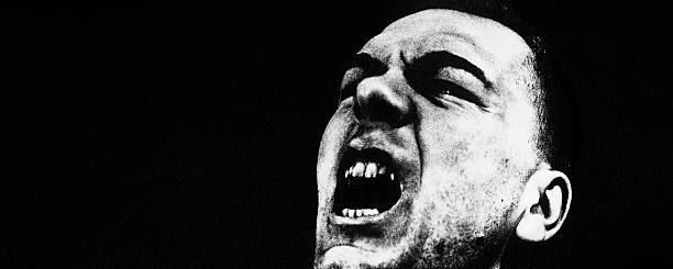High-contrast black and white portrait of a man with an open mouth shouting or yelling. Tight framing, dark background, and dramatic lighting visualize intense emotions such as anger, stress, frustration, or protest.