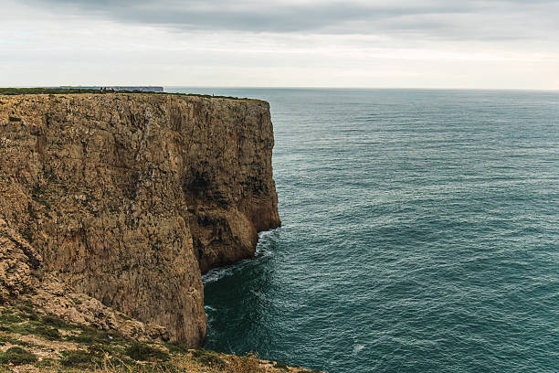 Steep vertical rock wall meets the calm sea under a soft overcast sky.