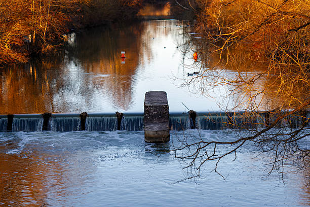 Autumn trees reflecting on flowing river water over a small weir at sunset