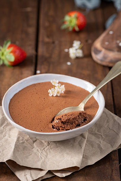 istock Velvety chocolate mousse in a white bowl, a golden spoon taking a bite, adorned with delicate white flowers, resting on crumpled parchment paper and a rustic wooden surface 2253859770