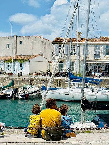 Vertical closeup photo of a man and has two children sitting at the edge of the harbour, looking out at boats moored on the water and apartment houses in the distance, St Martin de Re, Ile de Re.