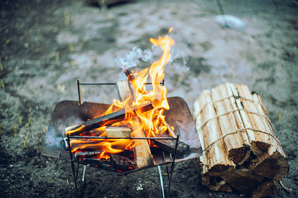 A campfire burning with wooden logs in a portable fire pit, with a bundle of firewood placed beside it. The scene represents outdoor camping, fire preparation, warmth, survival skills, and a calm natural lifestyle.