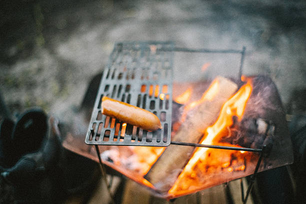 A single sausage being grilled on a metal grate over a campfire. Soft flames and glowing firewood create a calm and intimate outdoor cooking scene, representing simple camping meals, solitude, and a relaxed nature-focused lifestyle.