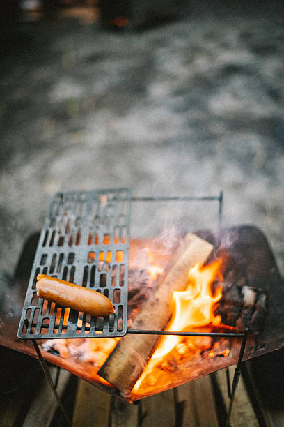 A single sausage being grilled on a metal grate over a campfire. Soft flames and glowing firewood create a calm and intimate outdoor cooking scene, representing simple camping meals, solitude, and a relaxed nature-focused lifestyle.