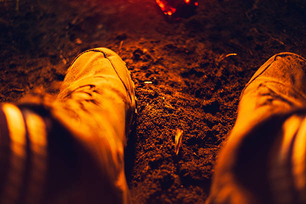 A first-person view of boots warmed by a campfire at night. The orange glow of the fire illuminates the ground and footwear, expressing warmth, comfort, relaxation, and a peaceful camping moment outdoors.