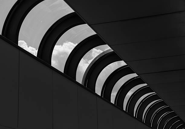 A monochrome architectural scene featuring a roof with distinctive half-round windows set against a dramatic cloudy sky. The black and white composition emphasizes contrast, texture, and geometric shapes, ideal for concepts of architecture, design, minimalism, and atmospheric photography.