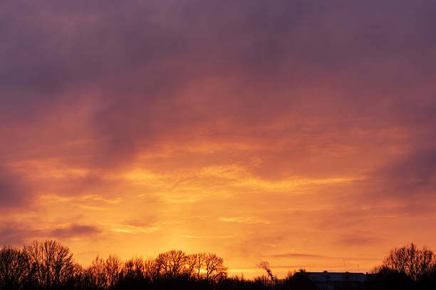Vivid warm sunset sky with orange and purple clouds illuminating the landscape, highlighting winter tree silhouettes and a distant building, creating a dramatic and atmospheric evening scene.