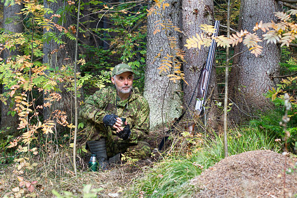 Autumn forest. The hunter is resting and drinking tea. His shotgun is nearby. There is an anthill in the foreground.
