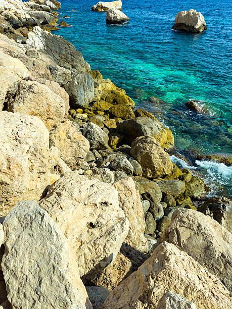 Bright sunlight illuminates the rugged rocks along the coastline, revealing turquoise waters that gently lap against the shore, creating a serene and inviting atmosphere.