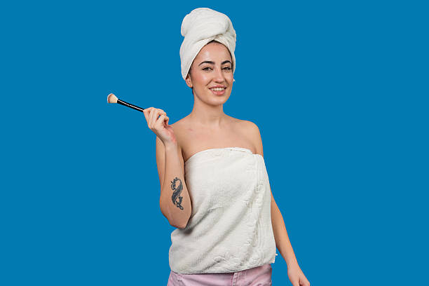 Woman standing with towel on head and body, holding makeup brush and smiling