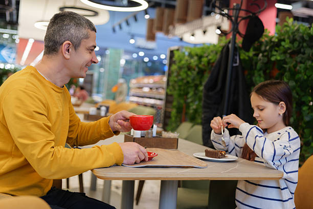 A man and a young girl share a delightful moment at a cafe, savoring cake and hot drinks. They are smiling and enjoying each other's company in a warm setting