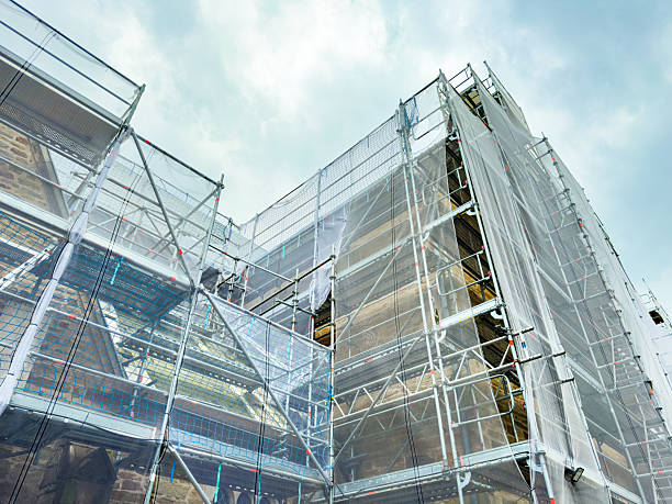 Workers carefully navigate the complex scaffolding as they restore an old structure, surrounded by protective mesh and the soft light of a cloudy afternoon.