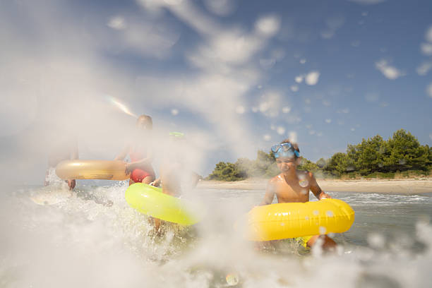 Captured from a unique surface-level perspective, this photo shows a group of teens joyfully throwing their youngest sibling into the air in a small northern Wisconsin lake. The child is caught mid-air, with water splashing around, making for a dynamic, playful moment. This action-packed scene, set under a blue sky on a sunny summer day, captures the carefree fun and excitement of youth, swimming, and family togetherness.