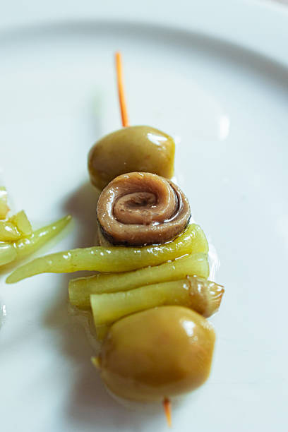 Gilda pintxo on a white plate containing olives, anchovy, and pickled peppers