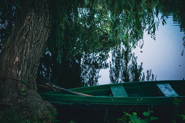 An old green boat is moored beside the quiet riverbank while a willow tree bends over the mirror-like water. Lush greenery surrounds the peaceful scene.