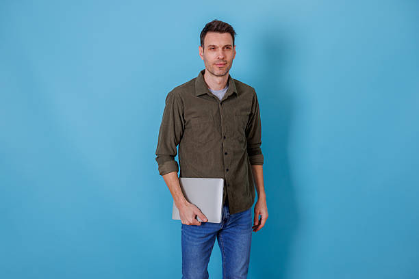 istock Young man standing holding a modern laptop against a blue background, representing technology and productivity 2253914004