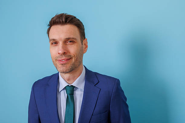 istock Professional man in a blue suit and teal tie smiling, looking confident against a plain blue background, representing business success 2253918756