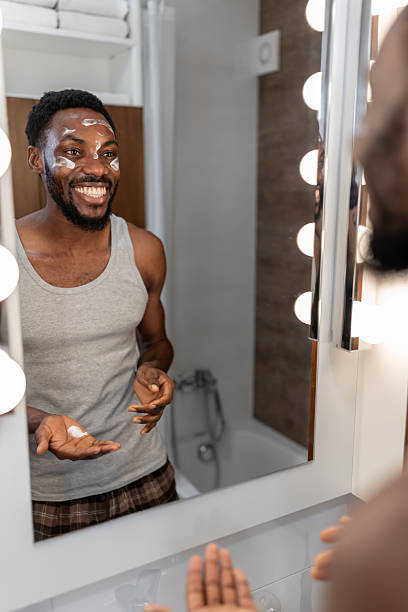 istock Smiling Black man applying face cream in a bathroom mirror, enjoying a simple skincare routine at home. Natural light, self-care moment, relaxed mood and positive emotion 2253921719