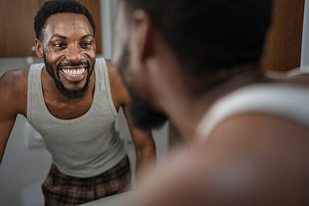 istock Cheerful Black man smiling at his reflection while touching his beard in the bathroom mirror. Relaxed home grooming moment, natural light, confidence and everyday self-care lifestyle 2253921892