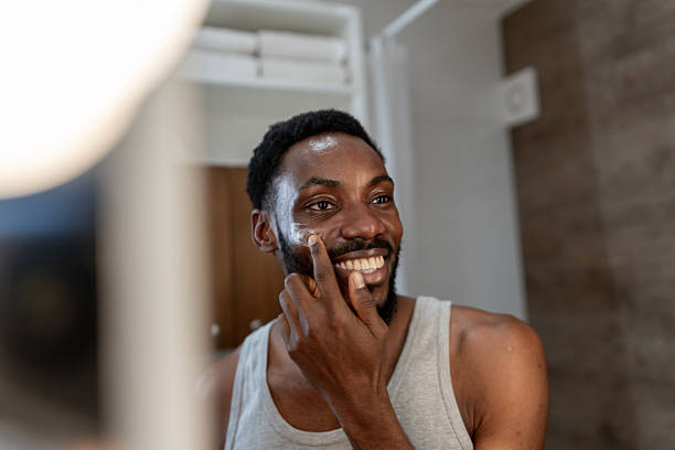 istock Smiling Black man applying face cream in a bathroom mirror, enjoying a simple skincare routine at home. Natural light, self-care moment, relaxed mood and positive emotion 2253921901