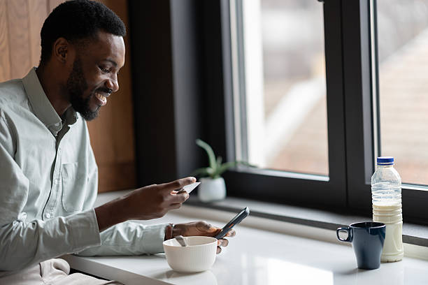 istock Smiling Black man at home during a morning breakfast makes an online payment on his smartphone while holding a credit card, representing digital banking, e-commerce, cashless payments, and a modern daily routine before work 2253922939