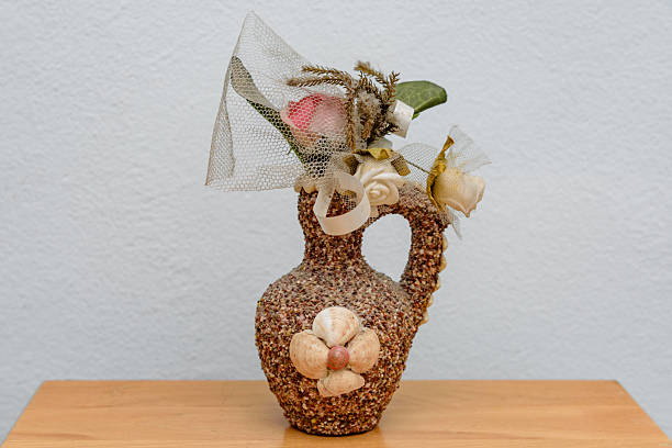 Handmade ceramic vase with a handle, covered in small seashells and containing artificial flowers.