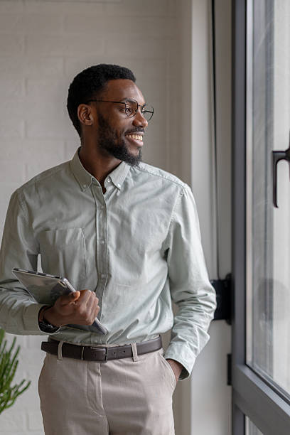 istock African American professional man holding a digital tablet while standing by an office window, thoughtful and focused, dressed in smart casual style in a modern workplace 2253923229