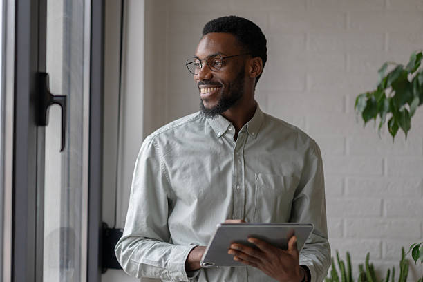 istock African American professional man holding a digital tablet while standing by an office window, thoughtful and focused, dressed in smart casual style in a modern workplace 2253923269