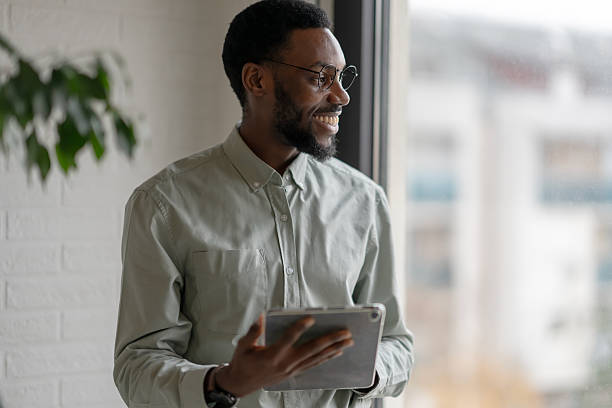 istock African American professional man holding a digital tablet while standing by an office window, thoughtful and focused, dressed in smart casual style in a modern workplace 2253923283