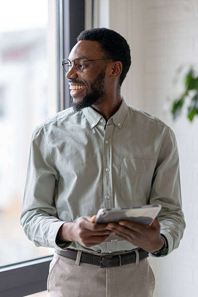 istock African American professional man holding a digital tablet while standing by an office window, thoughtful and focused, dressed in smart casual style in a modern workplace 2253923385