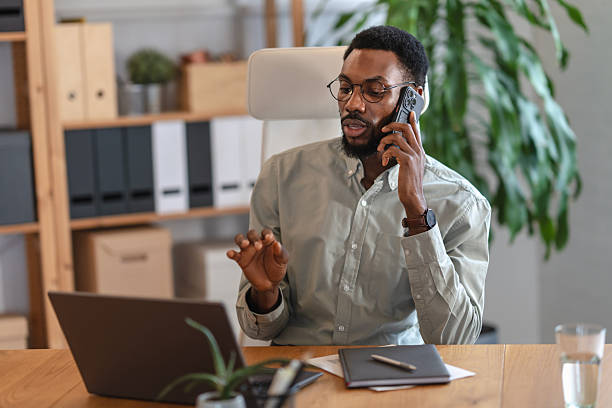 istock Confident Black man wearing glasses talks on a smartphone while working on a laptop in a modern home office, expressing focus, professionalism, multitasking, and calm control in a remote work setting 2253923681