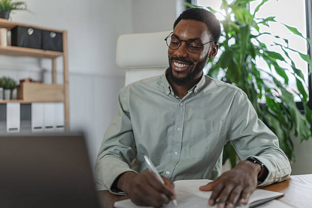 istock Smiling African American man working from home, writing notes at a desk with a laptop in a bright modern home office, relaxed and confident, focused on productivity, remote work, and business tasks 2253923716