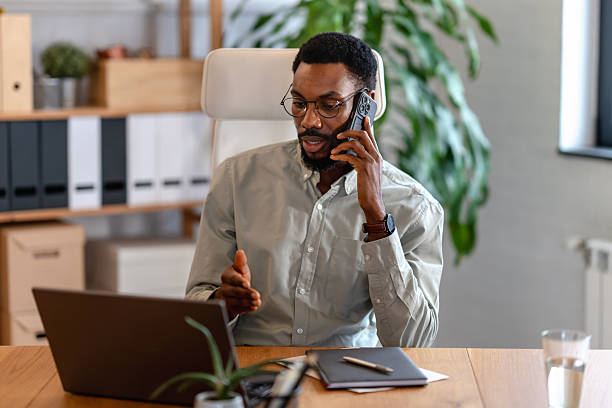 istock Confident Black man wearing glasses talks on a smartphone while working on a laptop in a modern home office, expressing focus, professionalism, multitasking, and calm control in a remote work setting 2253923726