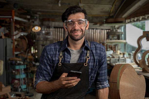 istock Smiling Carpenter in Workshop Using Smartphone, Craftsman Wearing Safety Goggles and Apron 2253924696