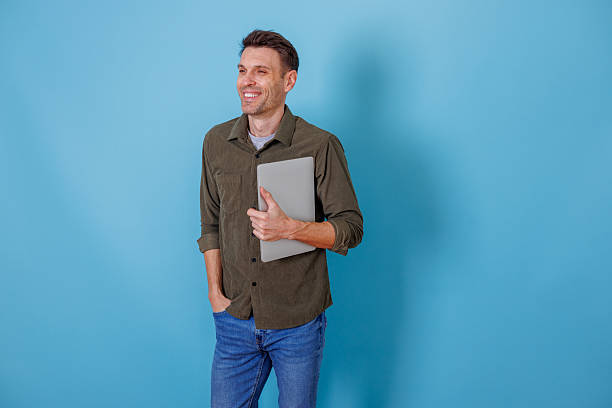 istock Young man holding laptop and smiling while standing against blue background, representing entrepreneurship and success 2253930891