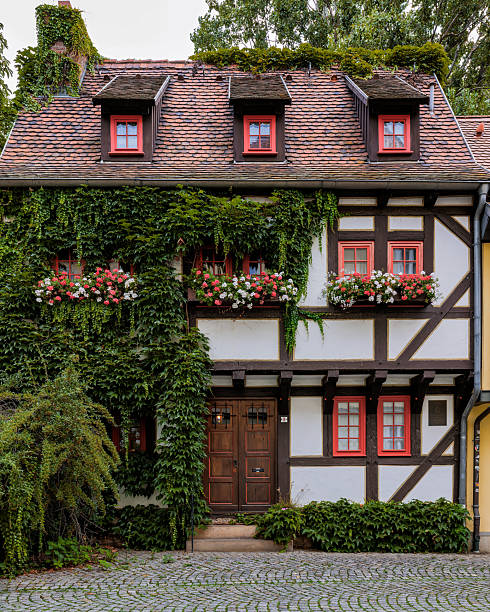 Located in a historic European town of Erfurt in Germany. The house features classic timber framing with dark wooden beams set against white plaster walls. The upper story slightly overhangs the ground floor, supported by visible wooden corbels.A steep, terracotta-tiled roof with three small dormer windows. Each dormer has a bright red frame that matches the windows on the lower floors.The windows are small, multi-paned, and painted a vibrant red, providing a striking contrast to the white and brown of the facade. The main entrance is a heavy, dark wooden double door located at the center, accessible by a small stone step.